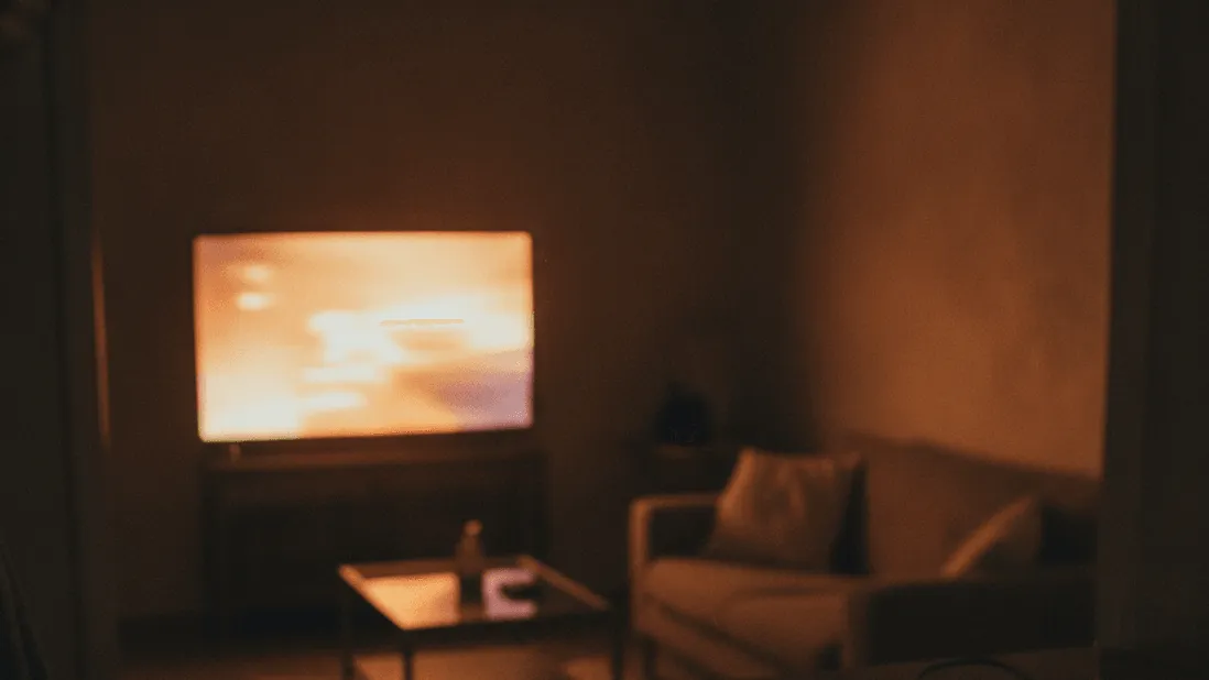 A person holds a tablet, its screen glowing in a dimly lit room with a television visible in the background.