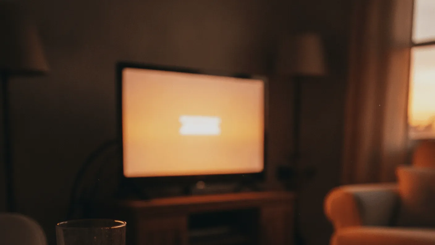A remote and a glass of water rest on a coffee table in a dimly lit living room with a television in the background.