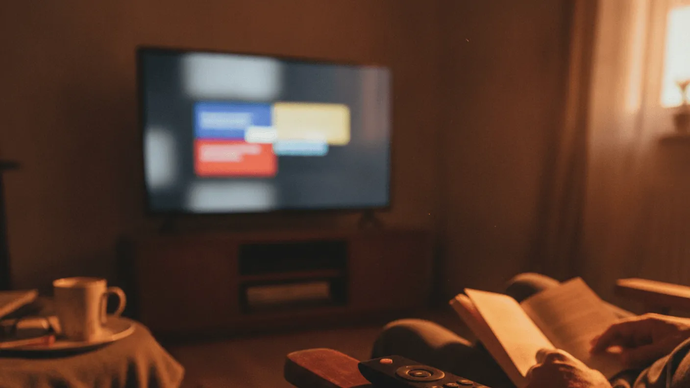 A television remote rests on a wooden armrest in a dimly lit room with a blurred screen in the background.