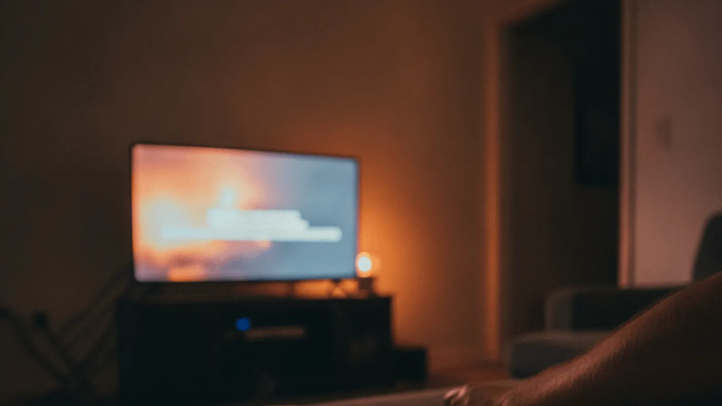 A remote control and coffee cup sit on a table, with a glowing television screen and a person's leg in the background.