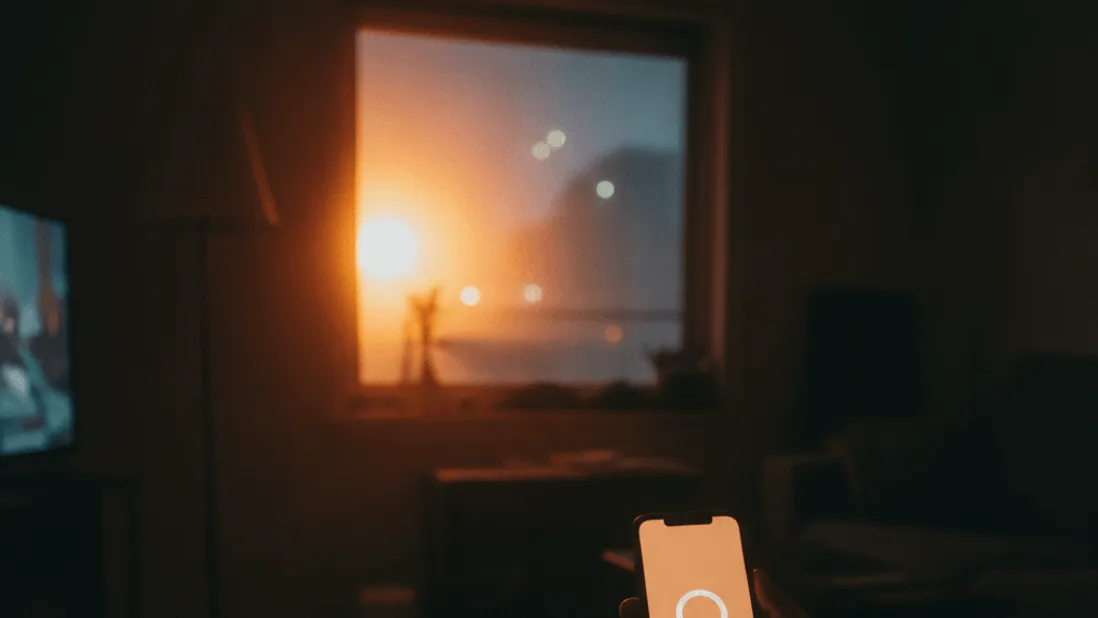 A hand holds a phone displaying a blank loading circle, on a couch armrest with a television glow reflecting on a window at dusk.