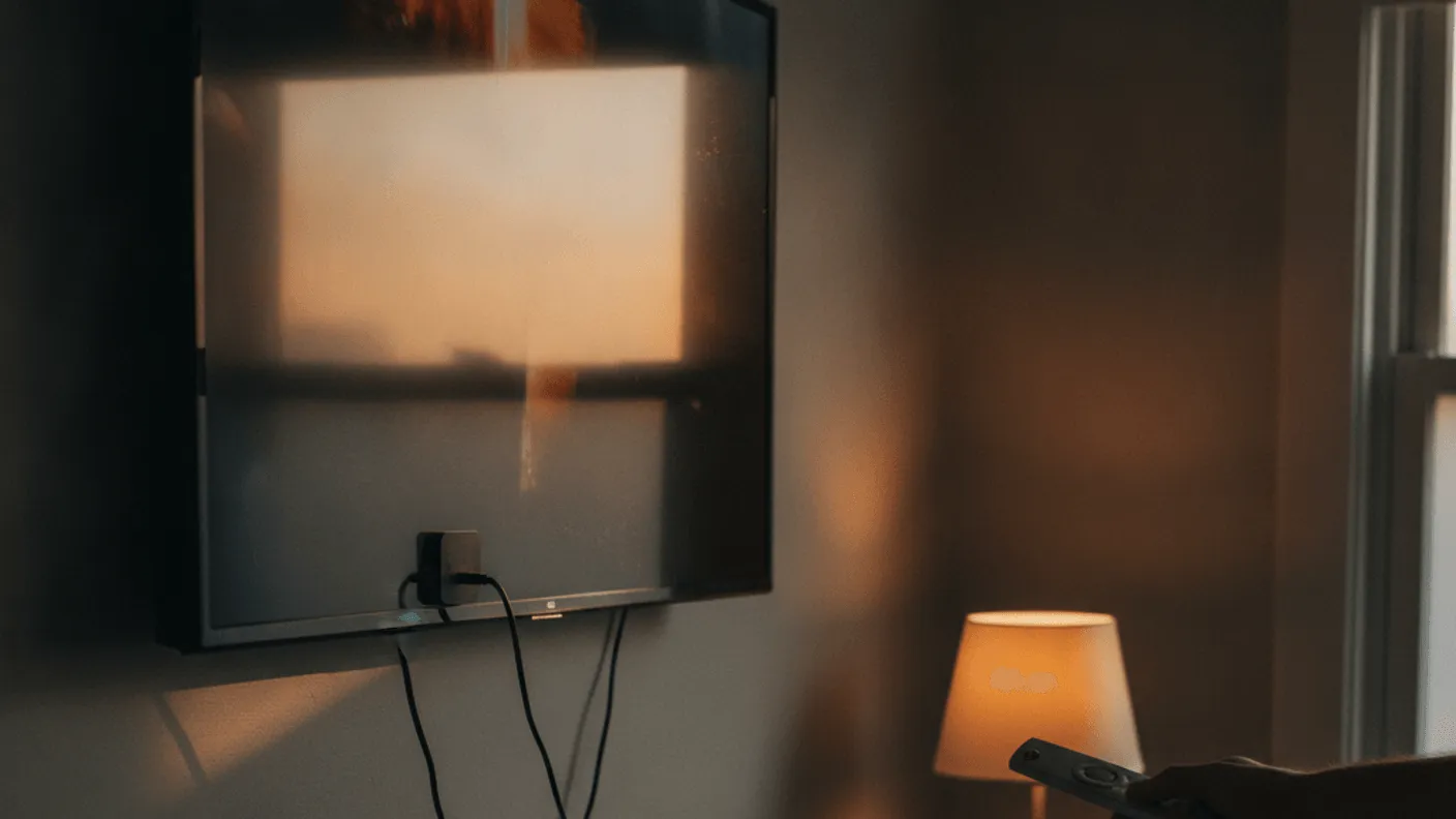 A small streaming dongle plugged into the rear ports of a wall-mounted television, with a person holding a remote.