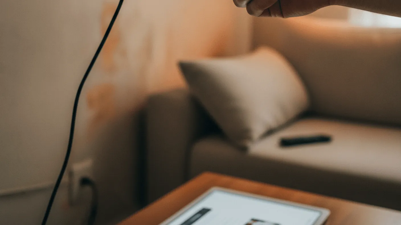 A hand inserts a streaming stick into the back of a wall-mounted television, with a couch and window in the background.