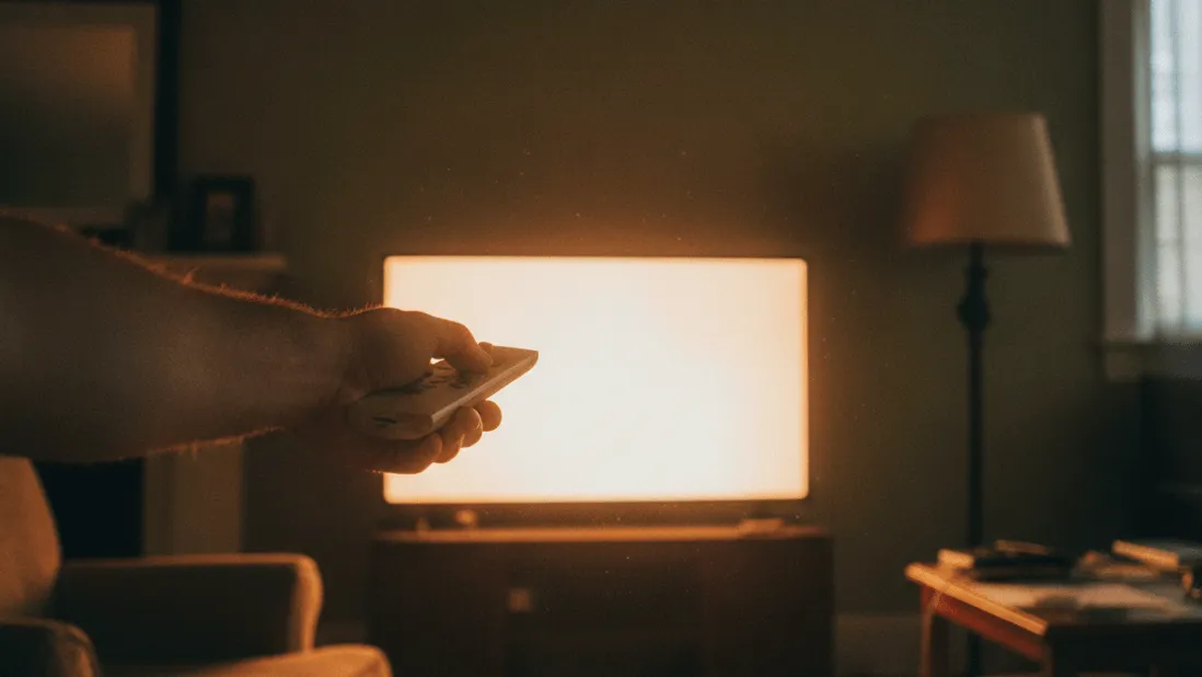 A man's hand holds a remote, pointed at a glowing television screen in a dimly lit living room.