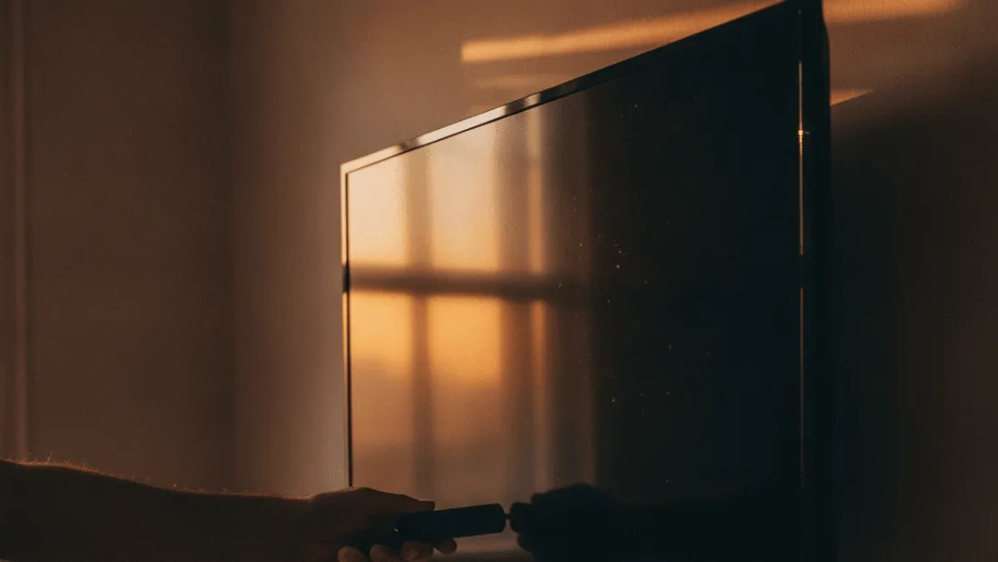A hand holds a streaming stick behind a wall-mounted TV with dramatic lighting from a window.