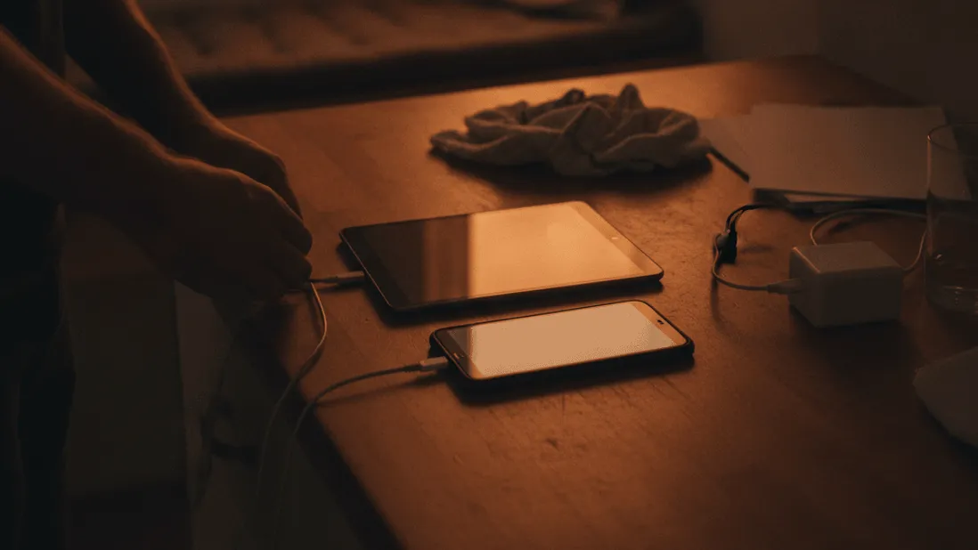 A man charges a tablet and smartphone on a kitchen counter in low, warm evening light.