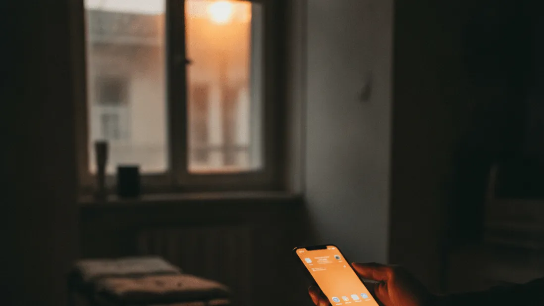 A hand holds a glowing smartphone above a kitchen counter next to a blank tablet at dusk.