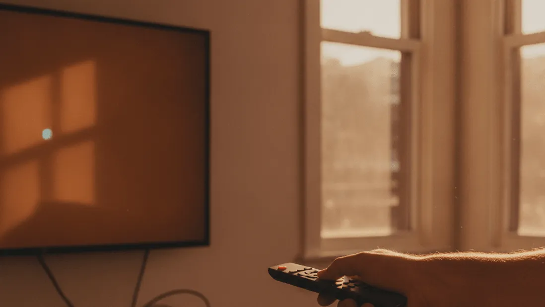 A hand points a remote at a media box beneath a television, with late afternoon light from a window.
